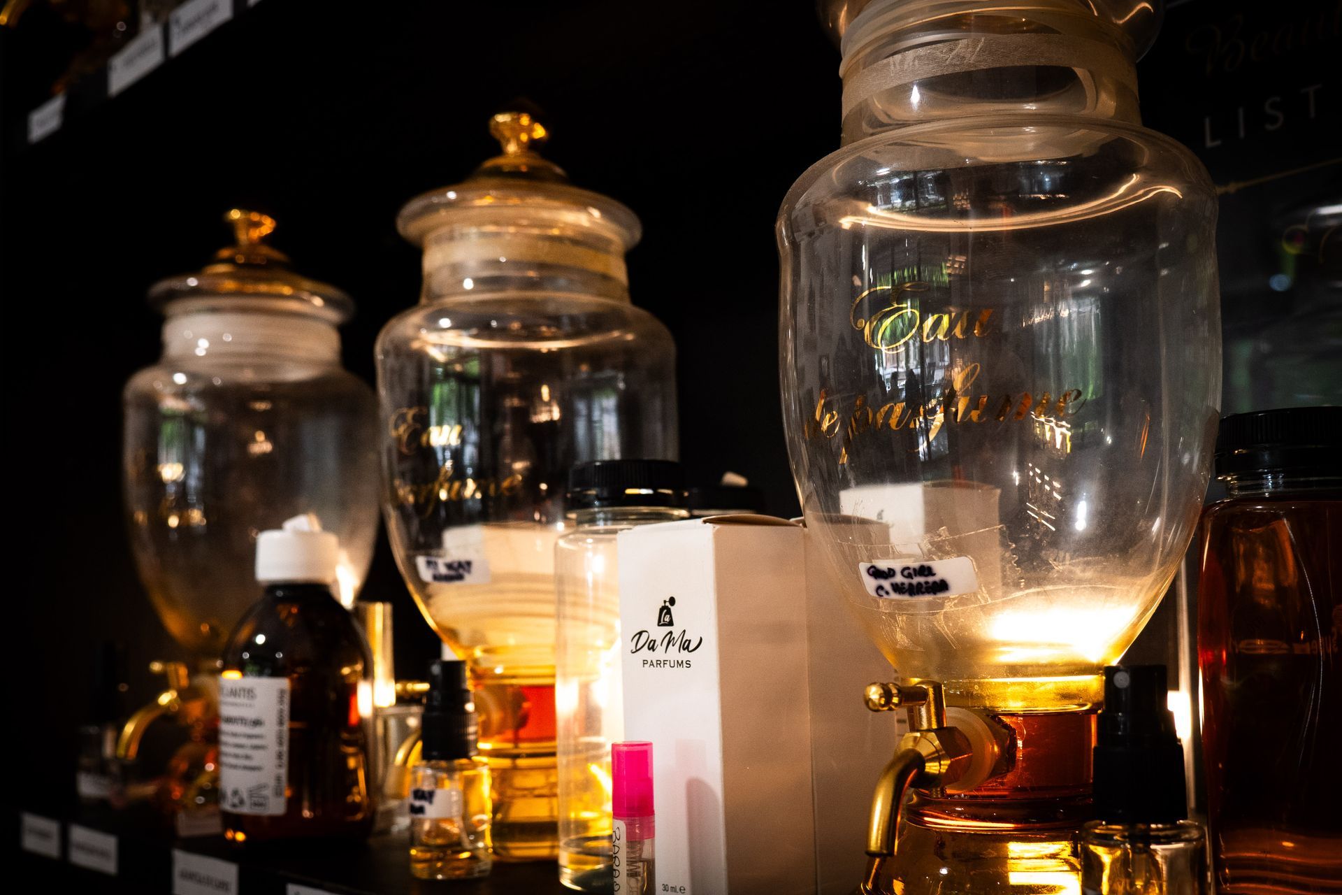 Tre dispenser di profumo in vetro con dettagli dorati, su uno scaffale con varie bottiglie di profumo.