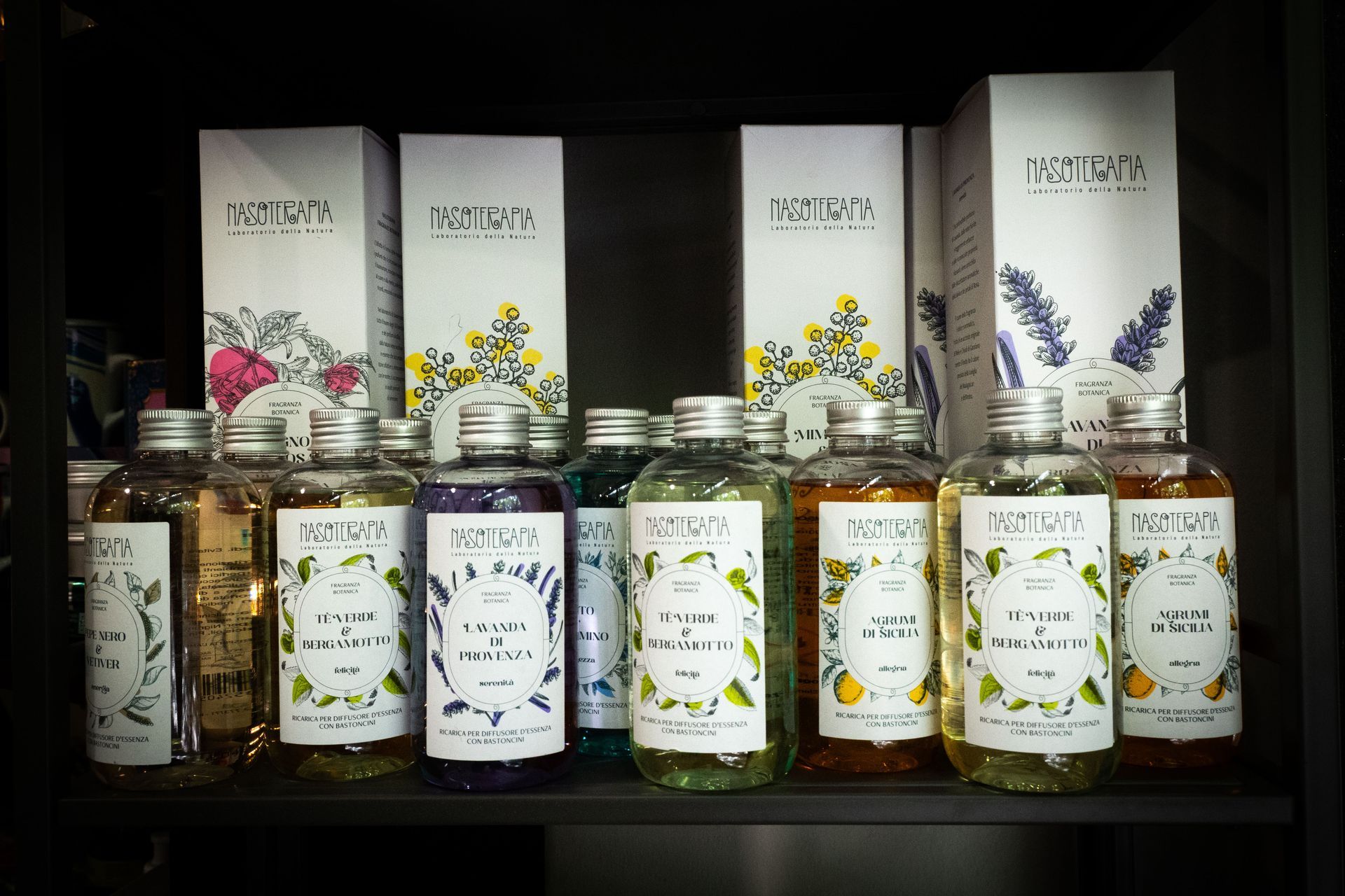 Bottiglie di oli profumati e le relative scatole disposte su uno scaffale, con illustrazioni botaniche ed etichette.