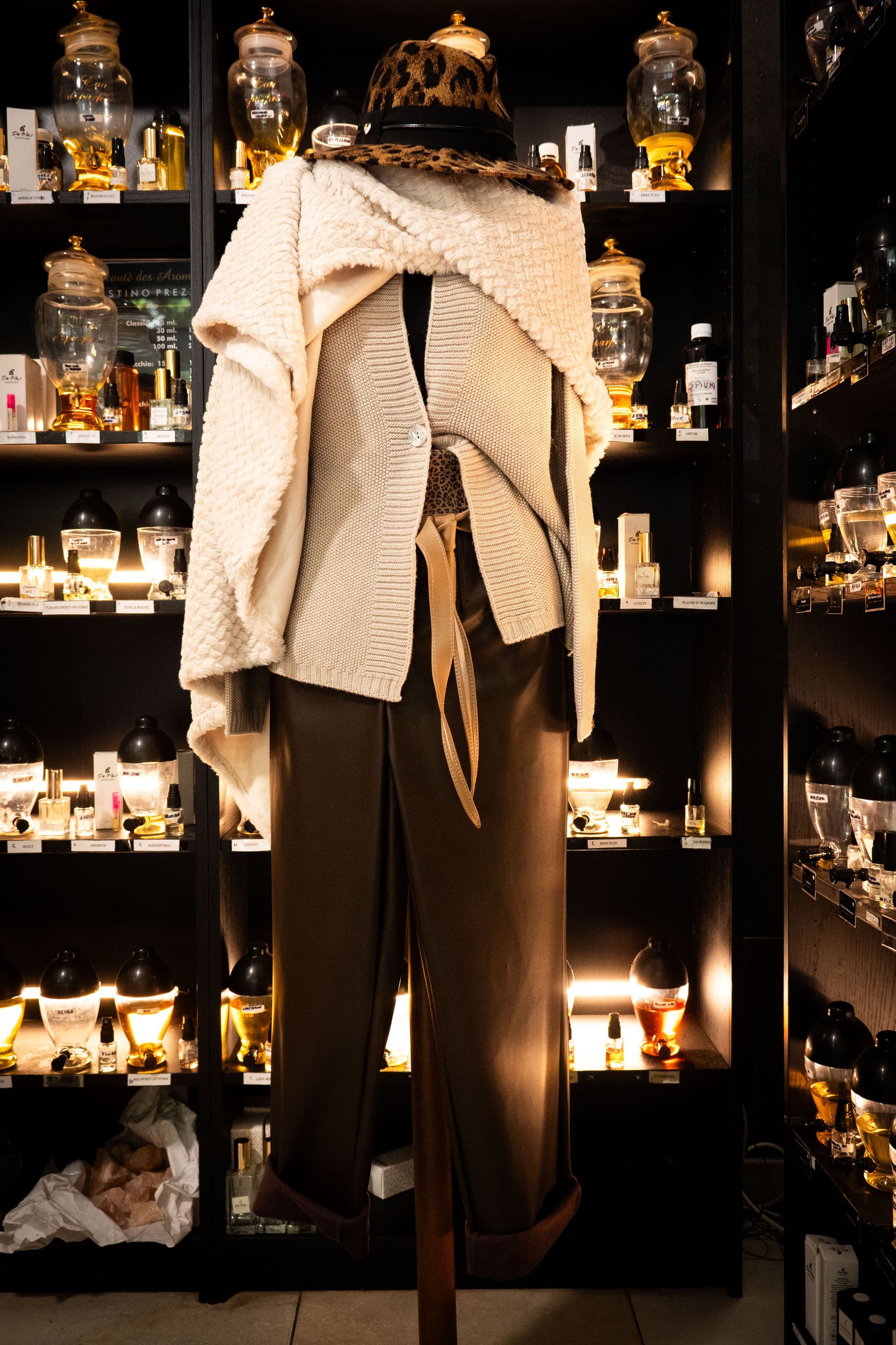 Manichino con top color crema, pantaloni marroni e cappello, in un negozio con bottiglie sugli scaffali.