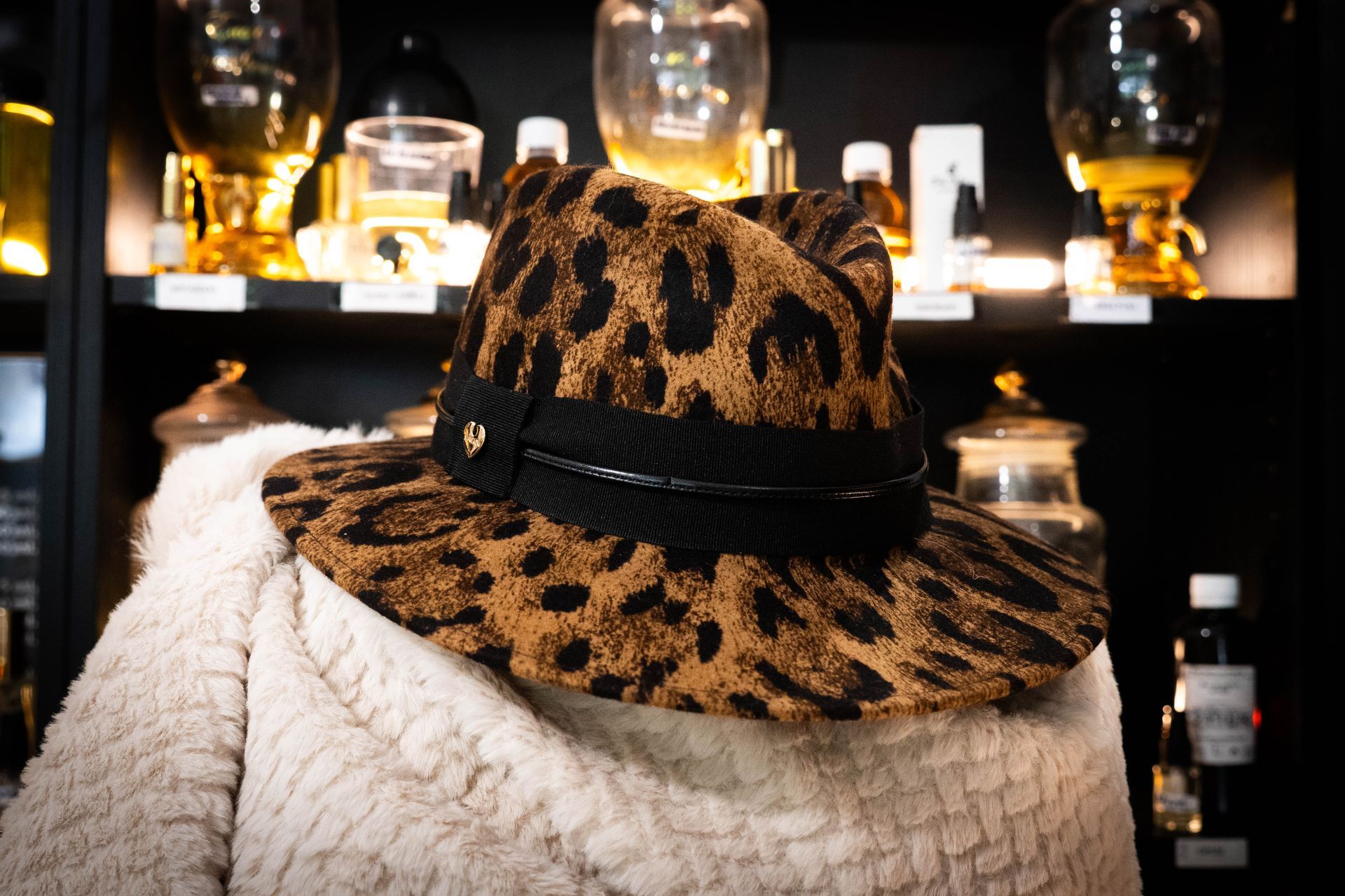 Cappello fedora con stampa leopardata e fascia nera, su un cappotto color crema davanti alle bottiglie di profumo.