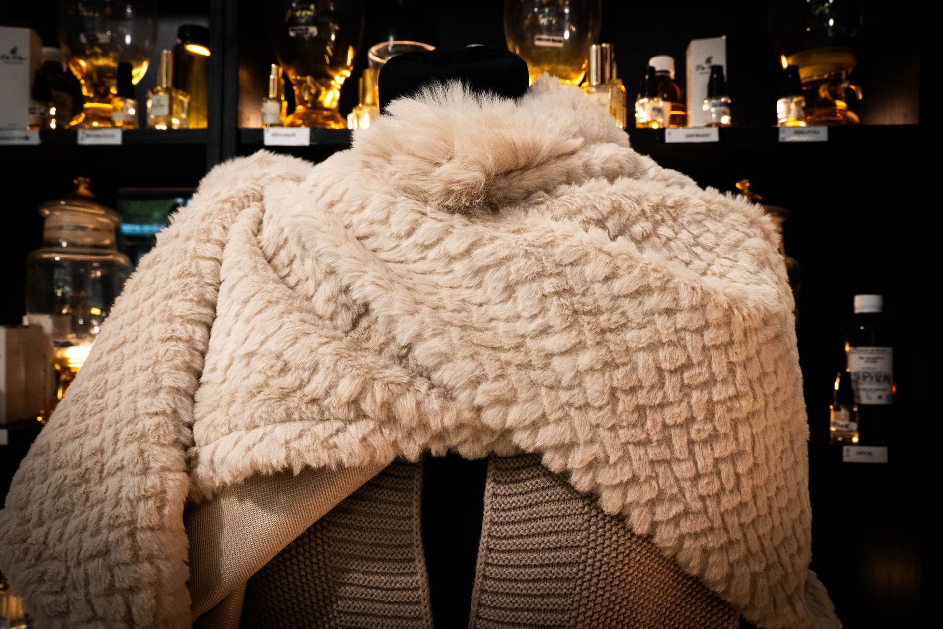 Uno scialle beige testurizzato è drappeggiato su un manichino. Sullo sfondo, scaffali con vari barattoli e bottiglie.