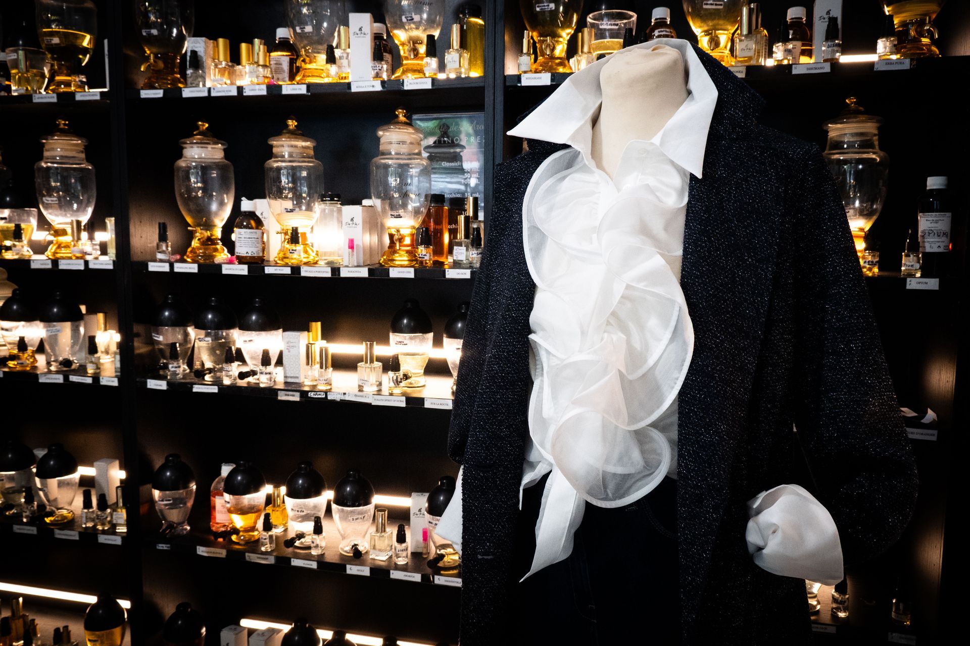 Manichino in giacca e camicia con volant, bottiglie di profumo sullo sfondo. Bianco e nero.
