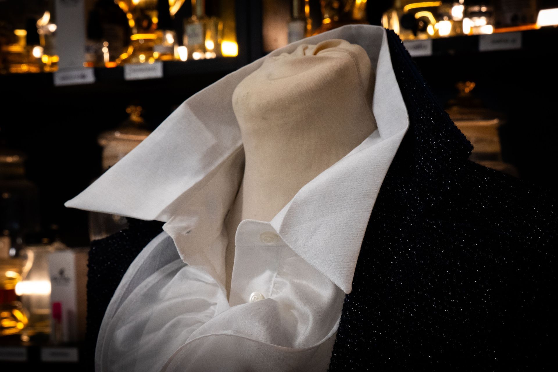 Testa e spalle di manichino con camicia bianca e giacca nera, davanti a scaffali pieni di barattoli.