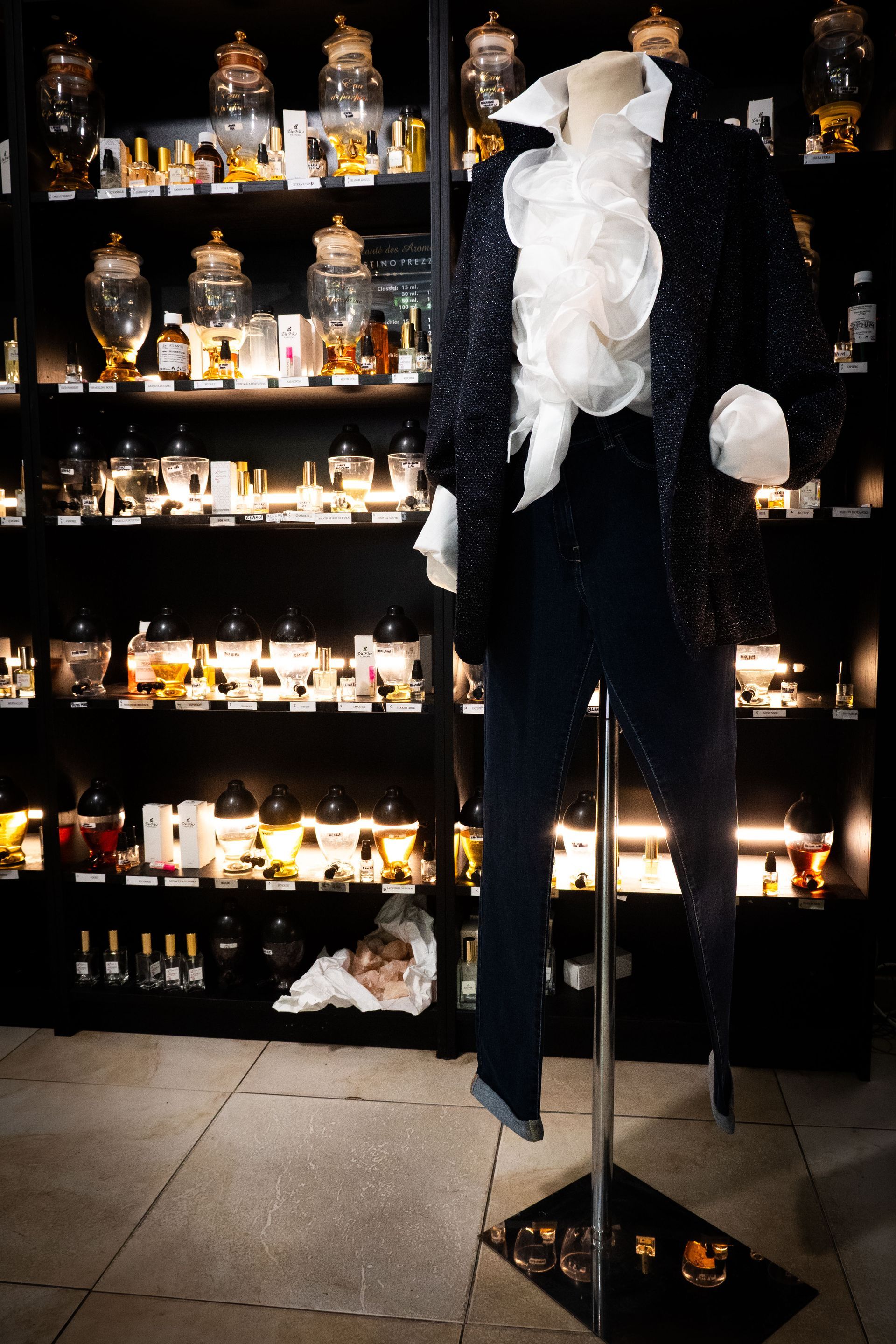 Un manichino vestito con una camicia arricciata e uno smoking davanti agli scaffali pieni di bottiglie di profumo.