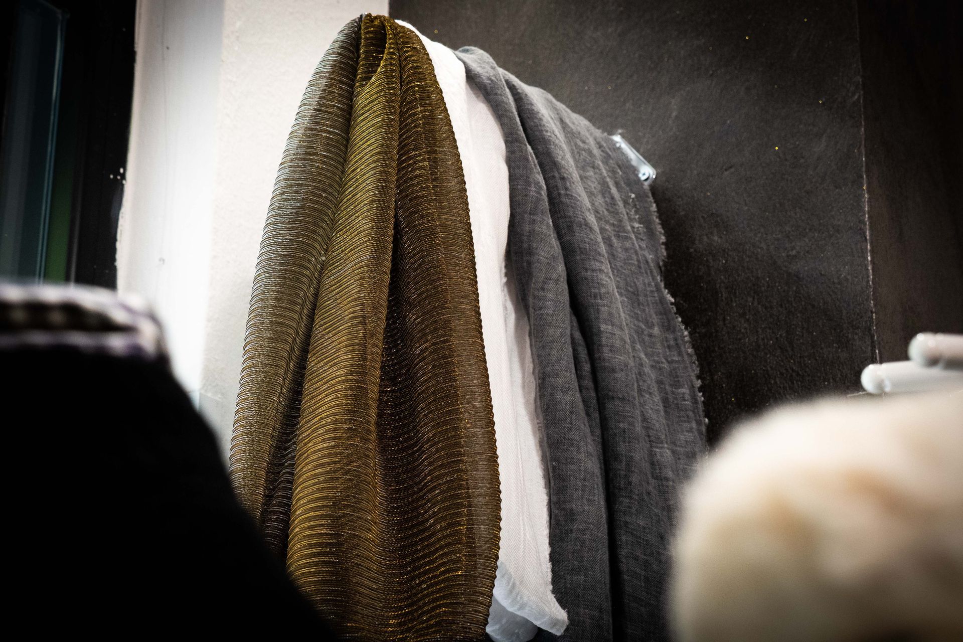 Tre tessuti drappeggiati: verde oliva, bianco e grigio, appesi a una parete scura.