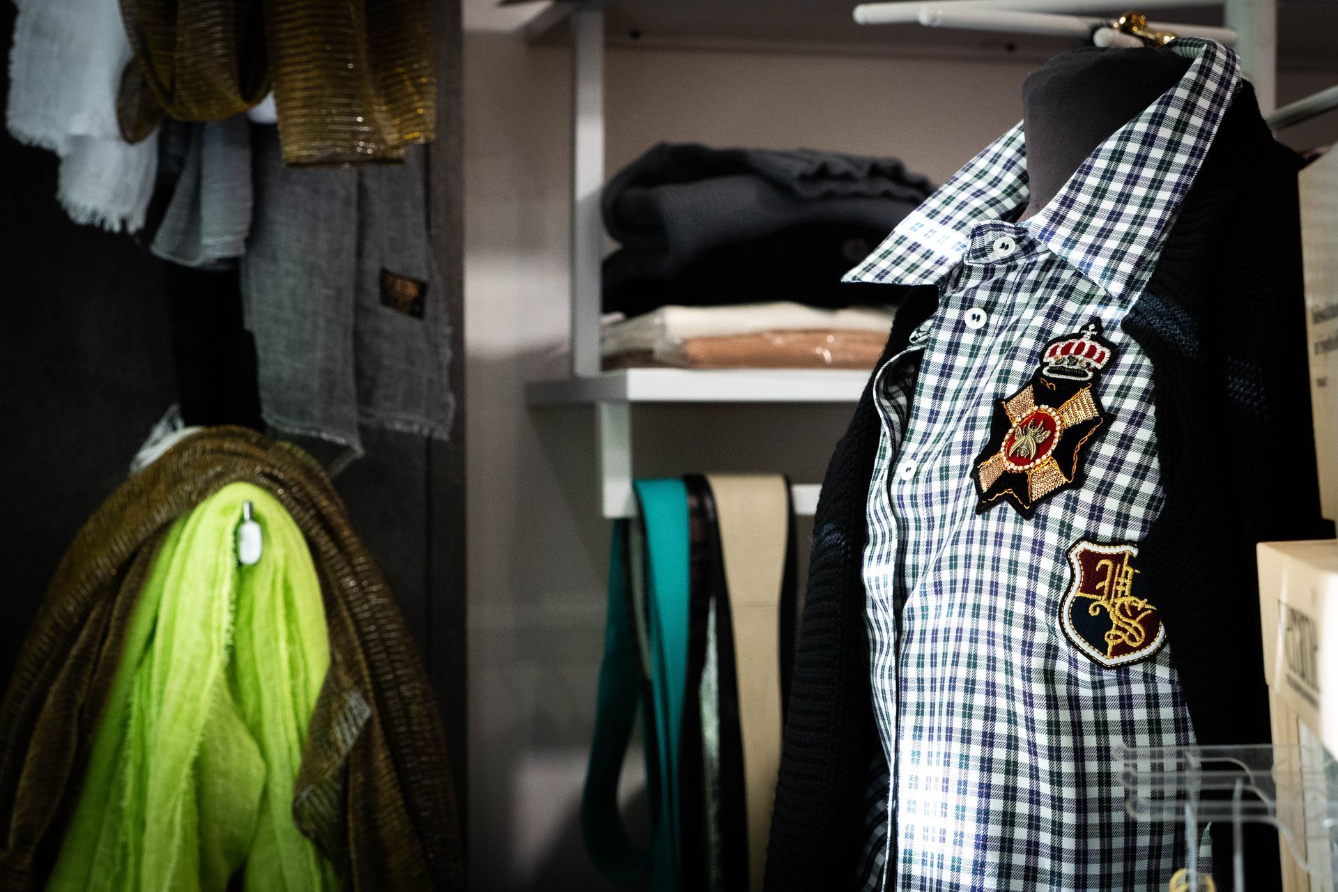 Esposizione di abiti: camicia a quadretti con toppe su un manichino, sciarpe e accessori in un armadio.