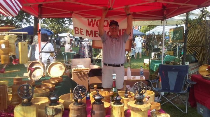 Man stands at wood craft stall, arms raised. Red canopy, wooden bowls, and carvings are displayed at an outdoor market.