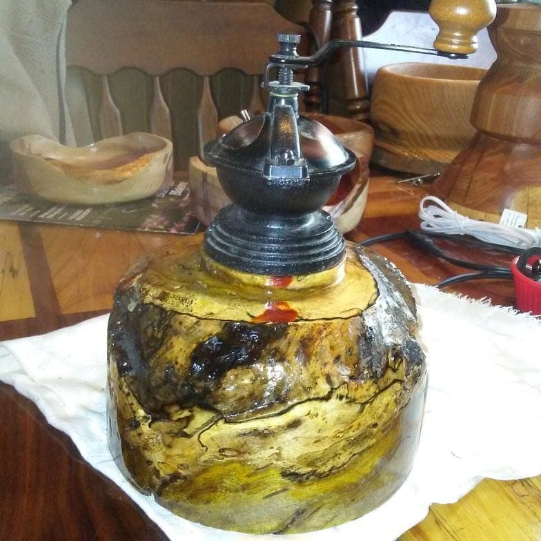 Antique coffee grinder atop a turned wooden base with a natural edge; yellow and brown tones.