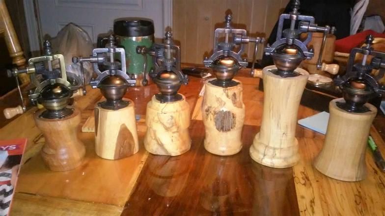 Six wooden and metal pepper grinders on a wooden table. Each has a unique wooden base.