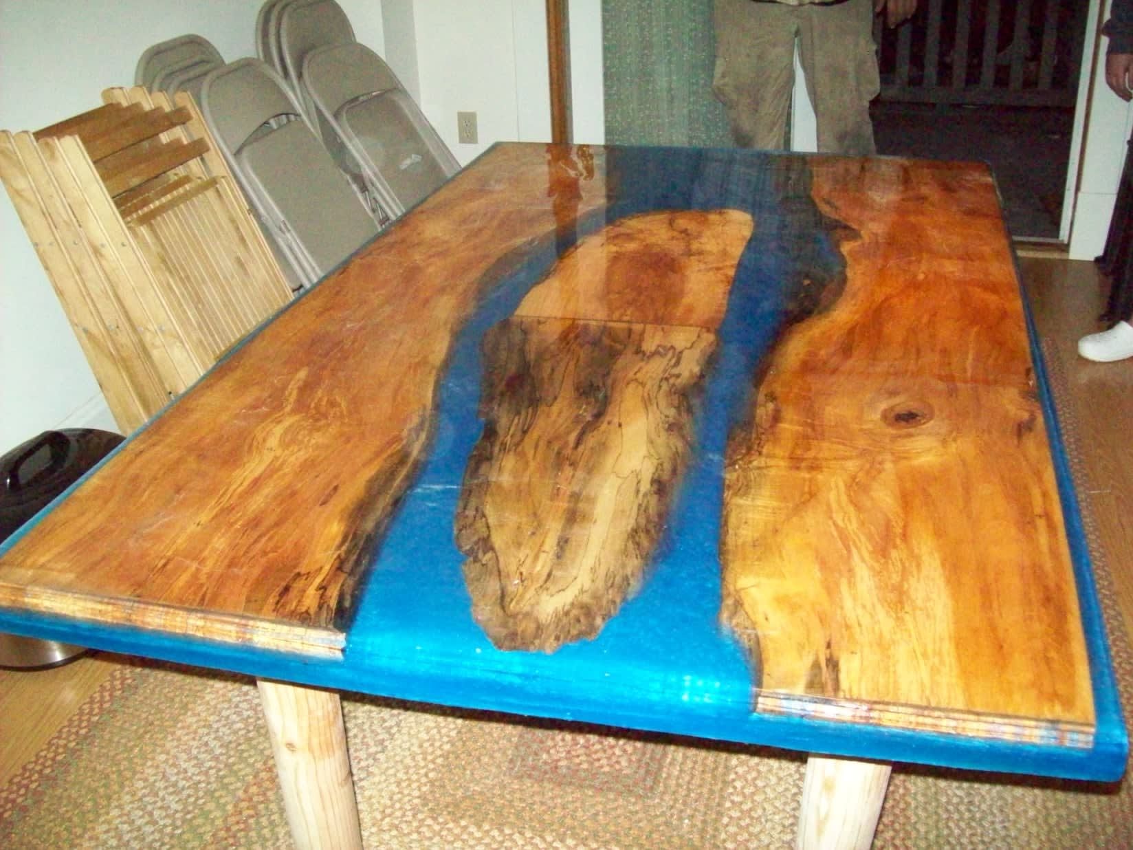 Wooden table with blue epoxy river, wooden legs, chairs and person in the background.