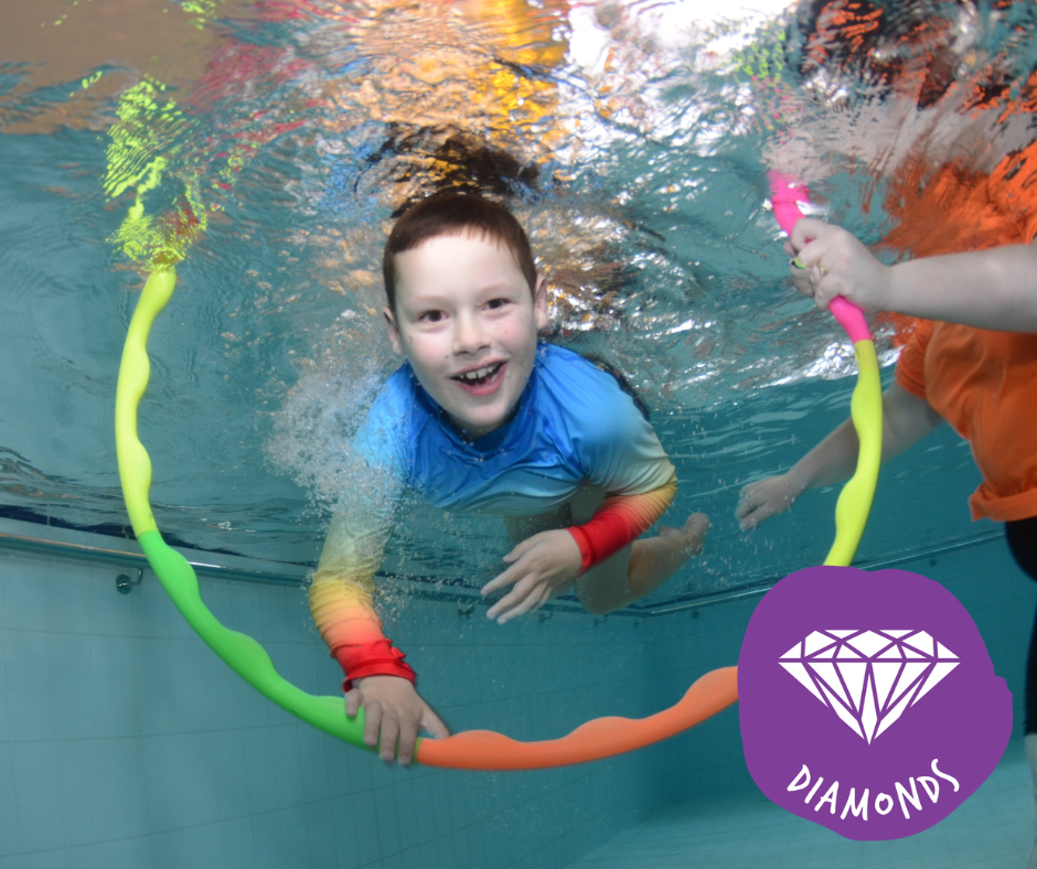 Baby in blue swim shirt, held by an adult in pool, laughing while reaching for toy.