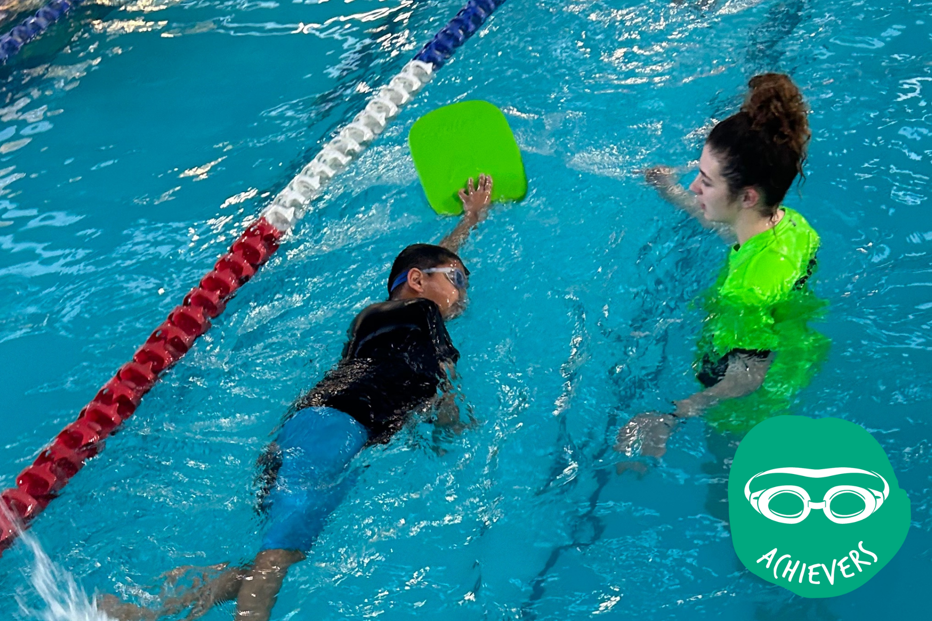 Child using a kickboard swimming, guided by an instructor in a pool.