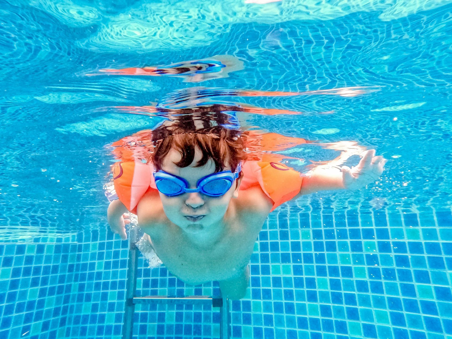 Boy with goggles and arm floats swims underwater in a blue tiled pool.