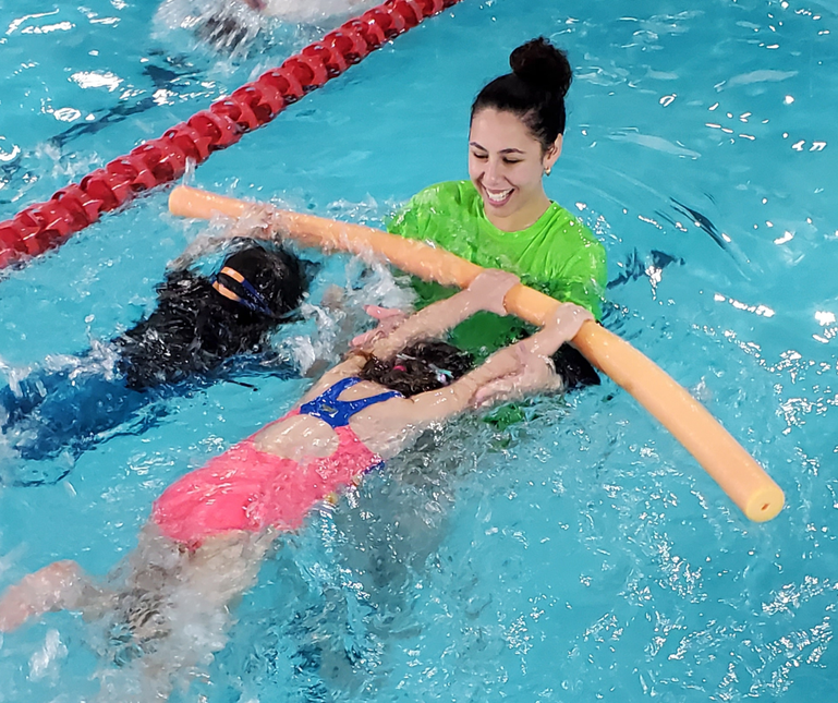 Woman in green shirt assists child learning to swim with a noodle in a pool.