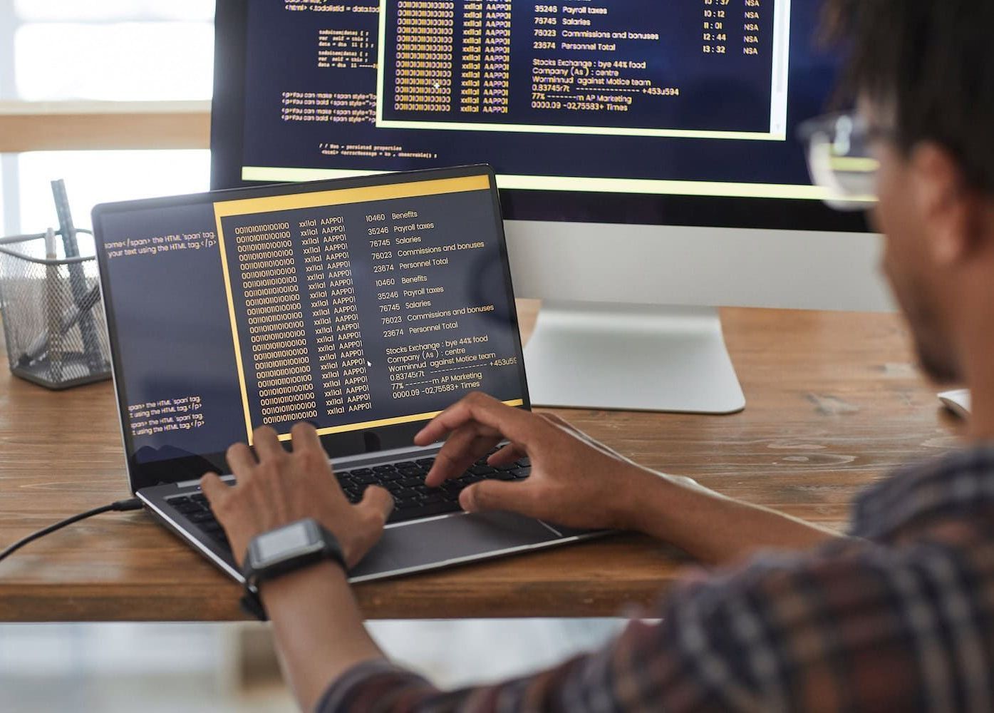 A man is typing on a laptop in front of a computer monitor