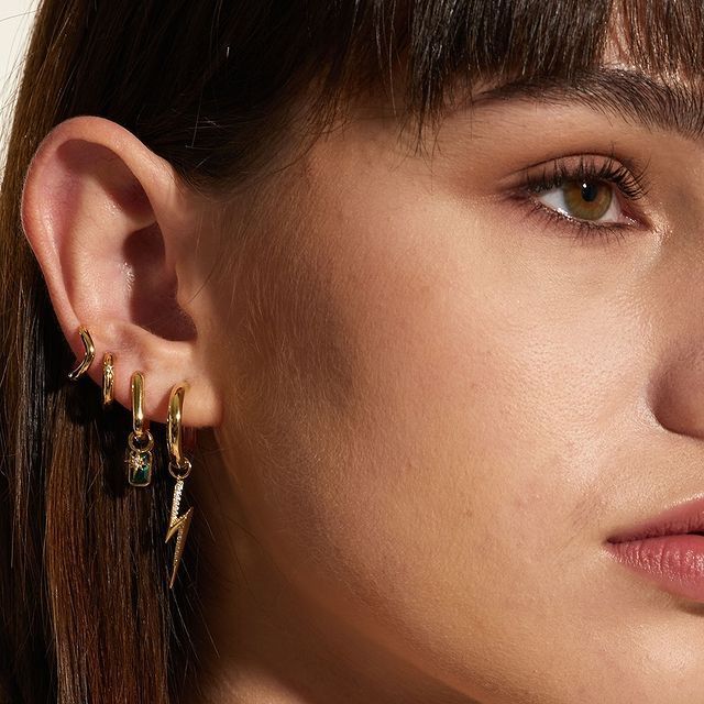 Oreille de femme avec plusieurs boucles d'oreilles en or : un éclair, une pierre précieuse et des créoles. Cheveux bruns, peau claire.