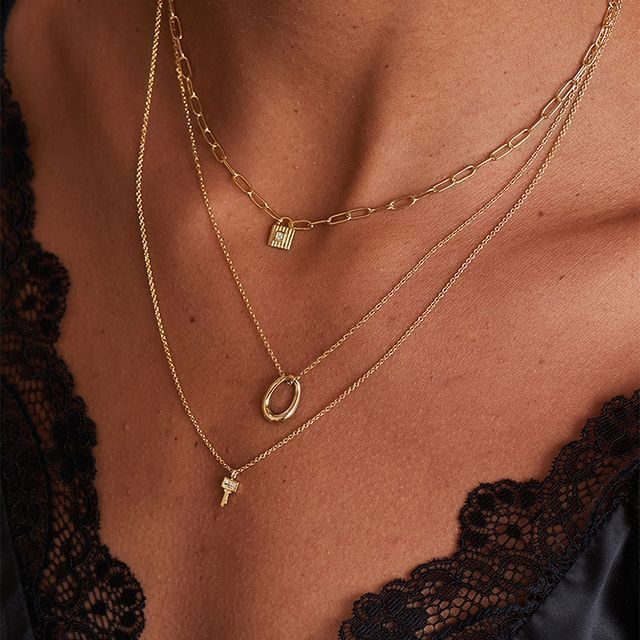 Gros plan d'une personne portant un collier en or superposé avec un pendentif en forme de cadenas, d'ovale et de clé.