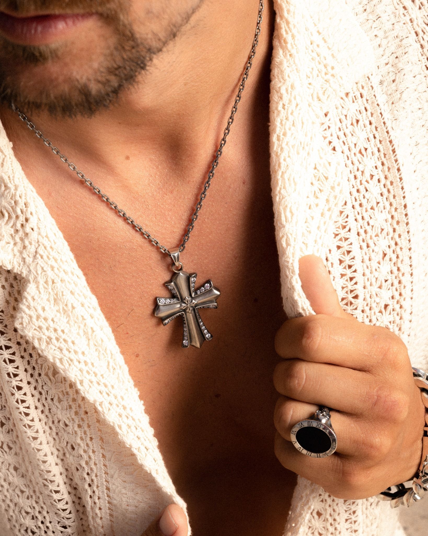 Un homme portant un collier en croix d'argent et une bague en pierre noire, tenant légèrement sa chemise ouverte.