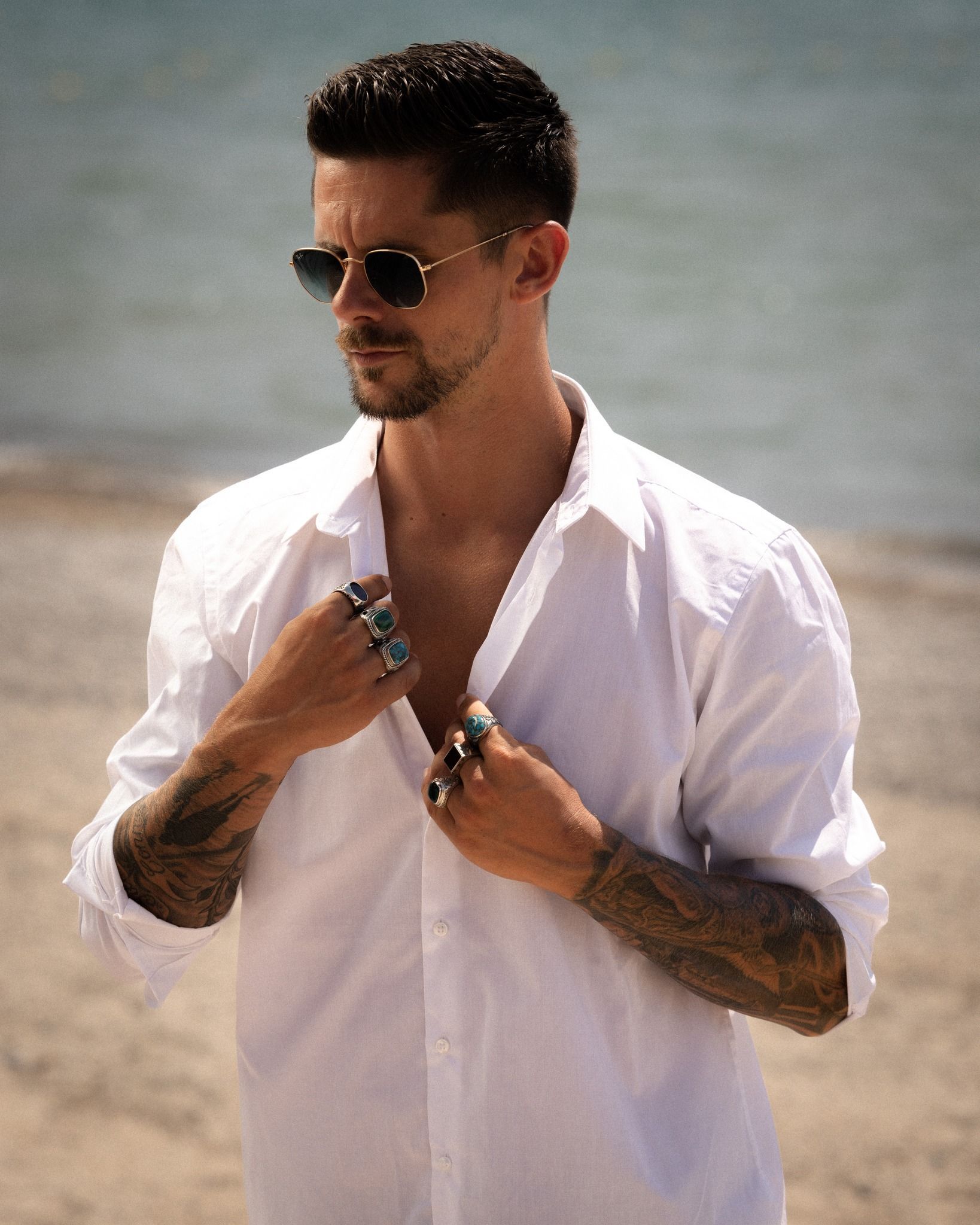 Un homme avec des tatouages, des lunettes de soleil et une chemise blanche sur une plage, déboutonnant sa chemise.