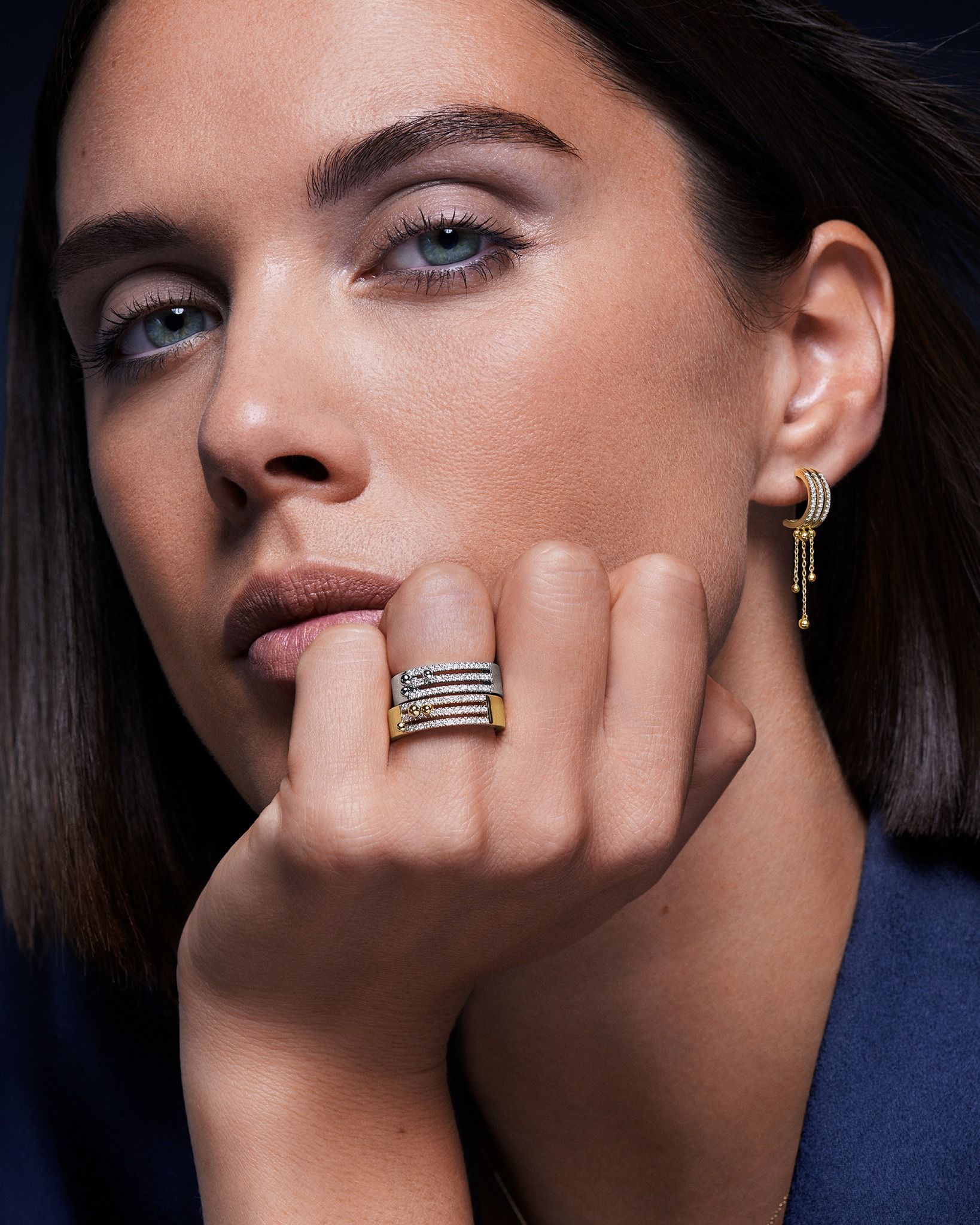 Femme aux yeux bleus, portant une bague et des boucles d'oreilles, le menton posé sur la main, sur un fond sombre.