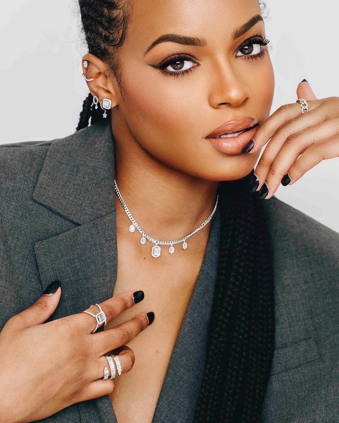 Femme en blazer gris avec bijoux, ongles noirs, eye-liner foncé.
