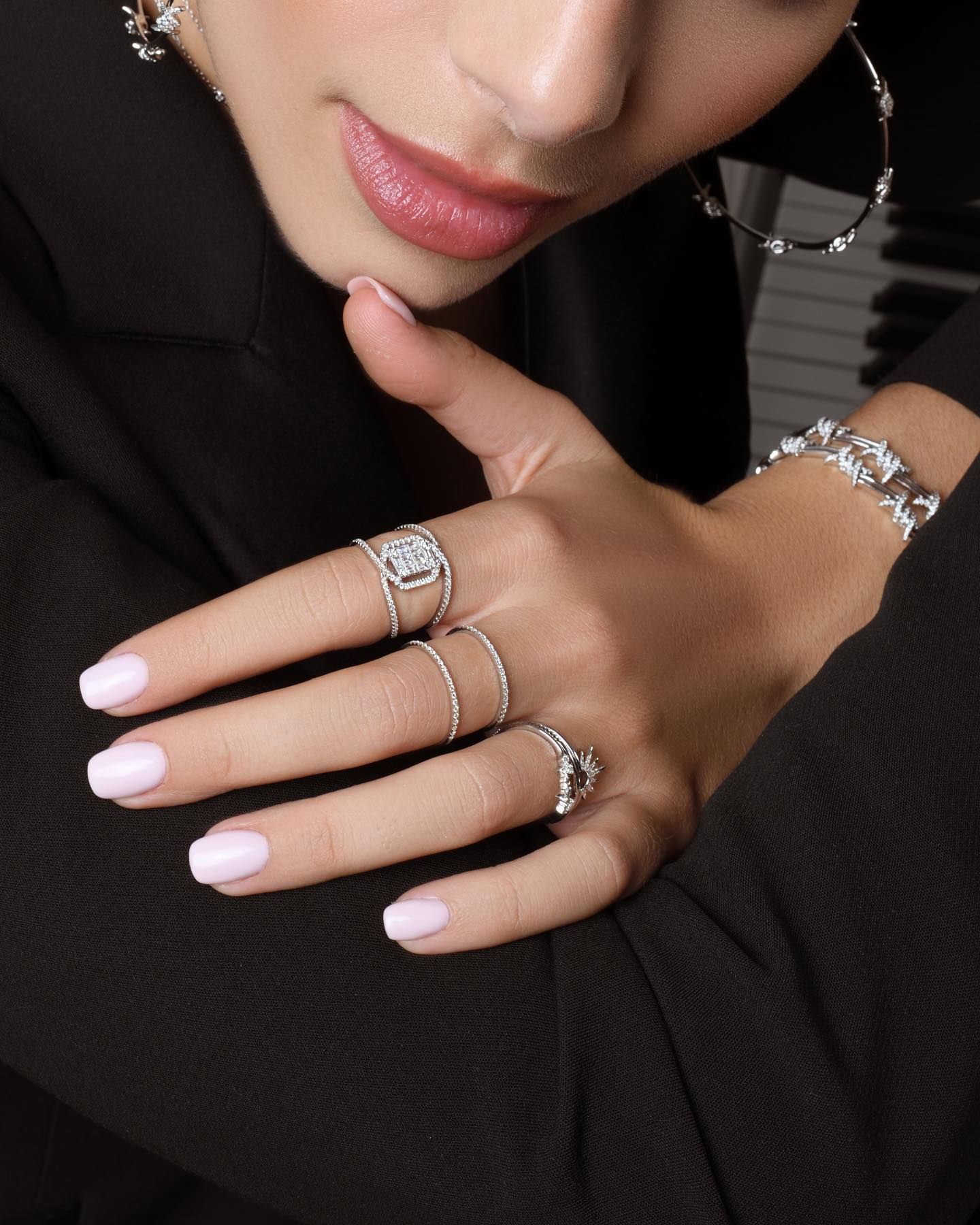 Main de femme portant des bagues, un bracelet et des créoles ; vernis à ongles rose et blazer noir.