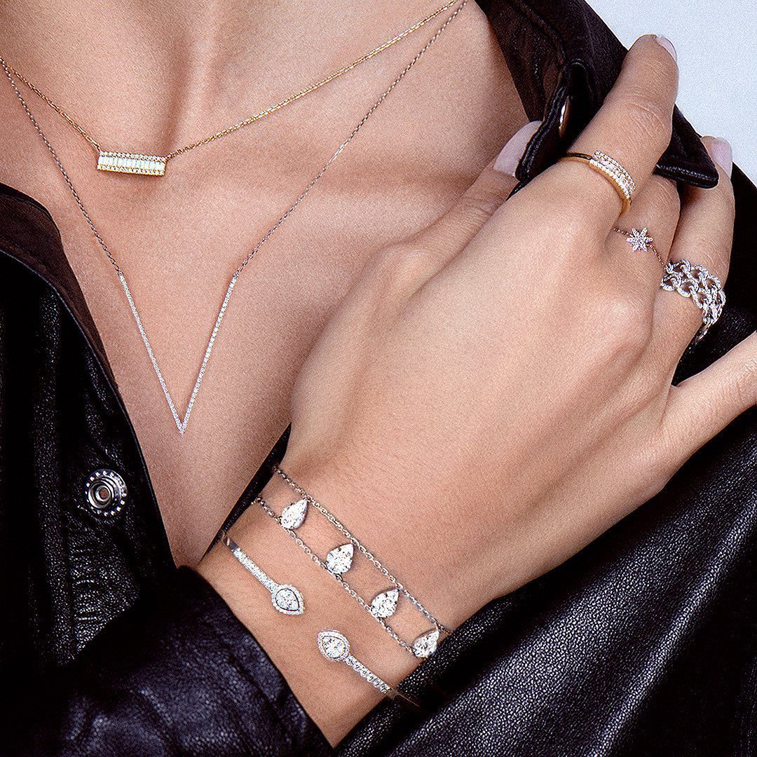 Main d'une femme portant des colliers, des bagues et des bracelets superposés, contre une veste en cuir noir.