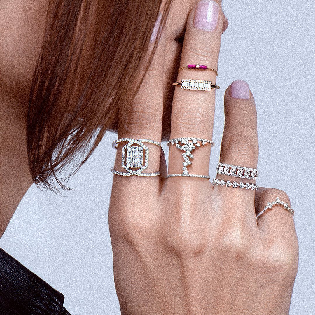 Main de femme avec plusieurs bagues en or et en diamant, doigts sur le visage, sur un fond clair.