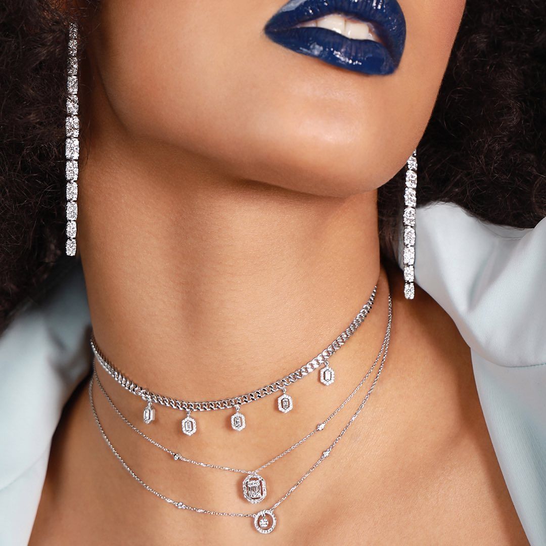 Femme portant des boucles d'oreilles et des colliers en diamant, du rouge à lèvres bleu foncé et une chemise bleu clair.