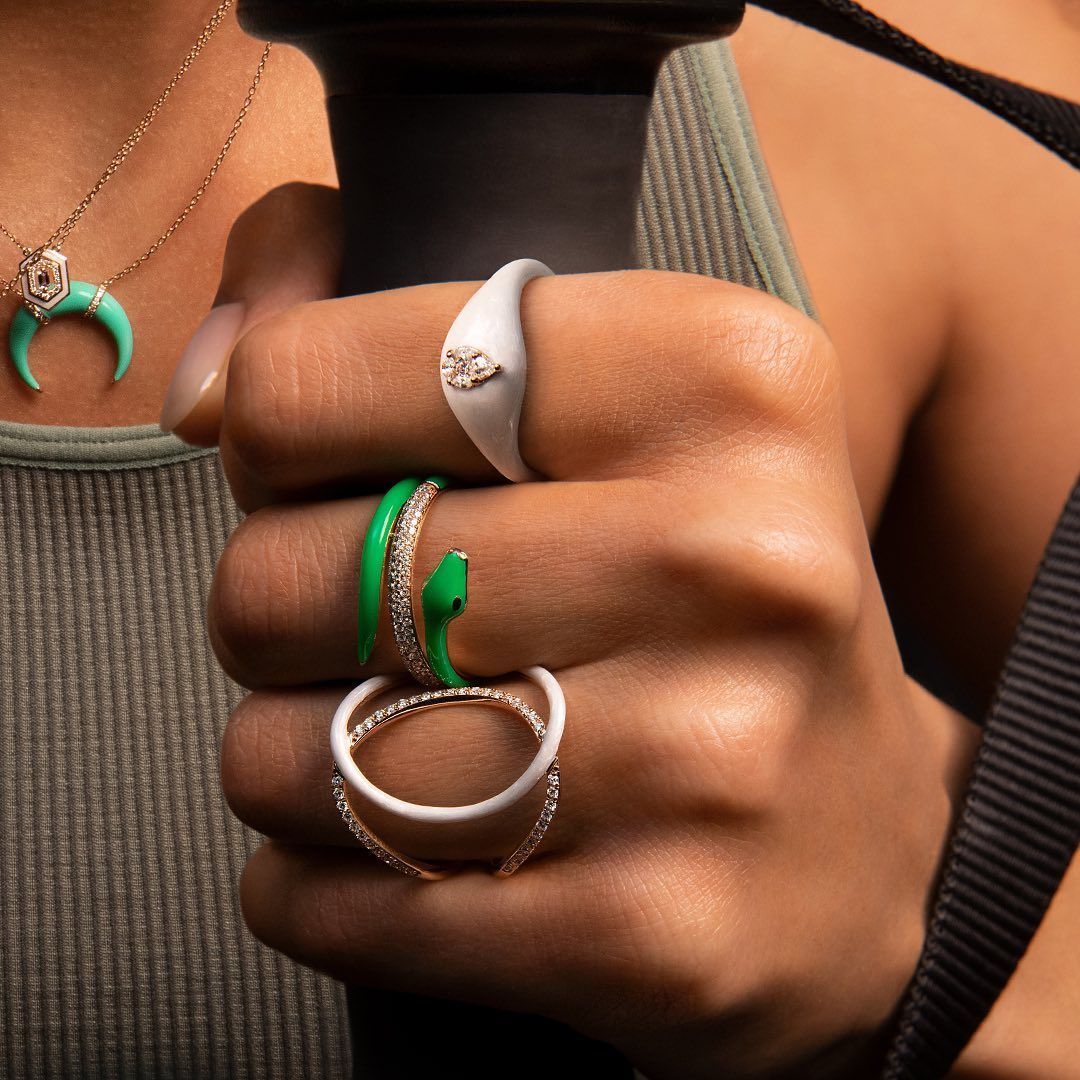 Main d'une personne avec plusieurs bagues aux doigts : émail blanc et vert, motif serpent, tenant un objet noir.