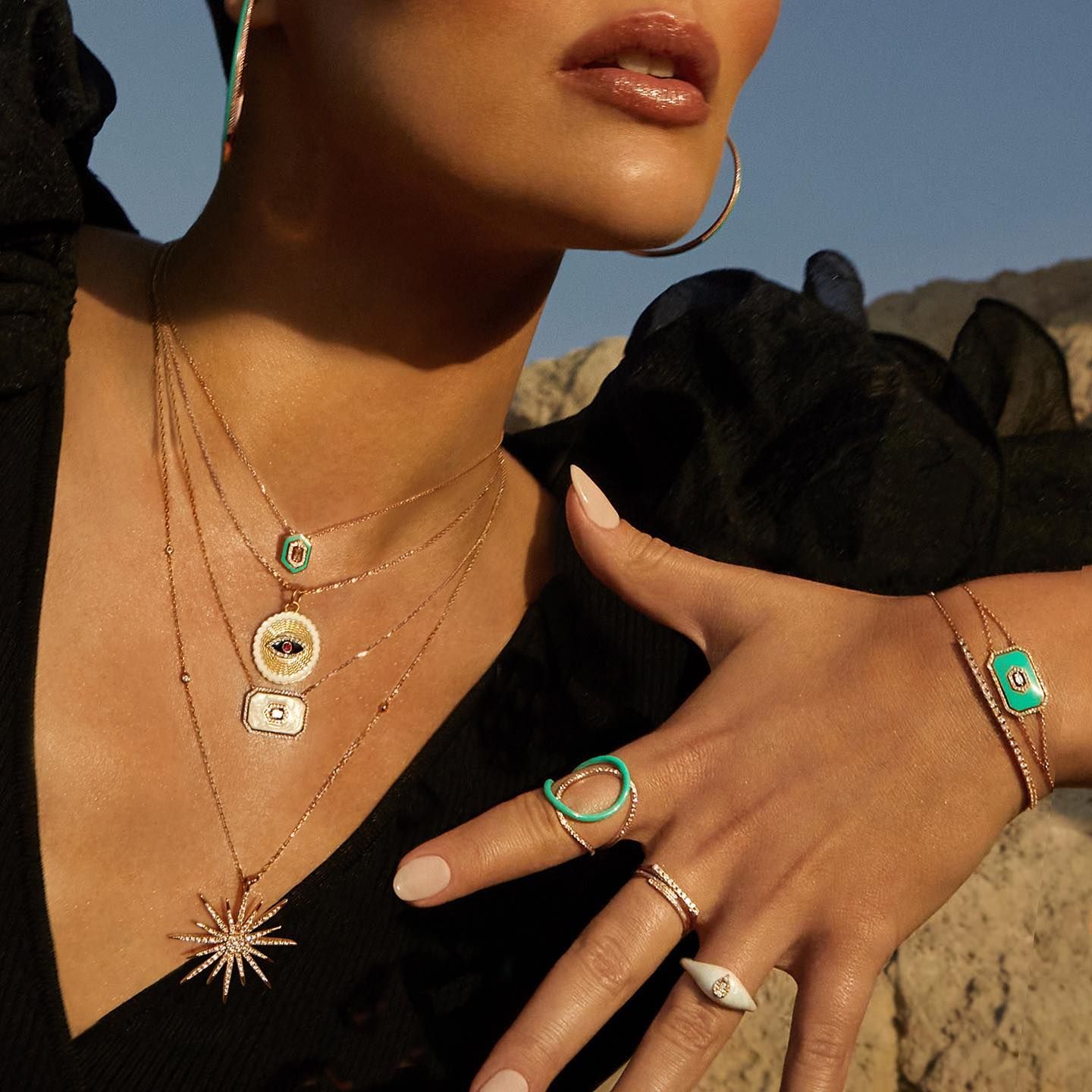 Femme portant des bijoux en or ; colliers, bagues et bracelet avec des accents de turquoise et de perles, près d'une plage rocheuse.