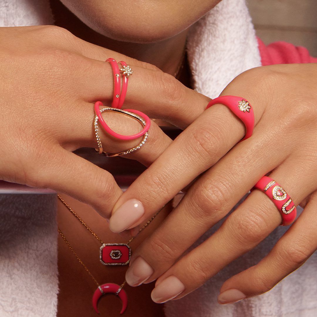 Mains de femme ornées d'anneaux rose vif et d'un collier, sur fond rose flou.