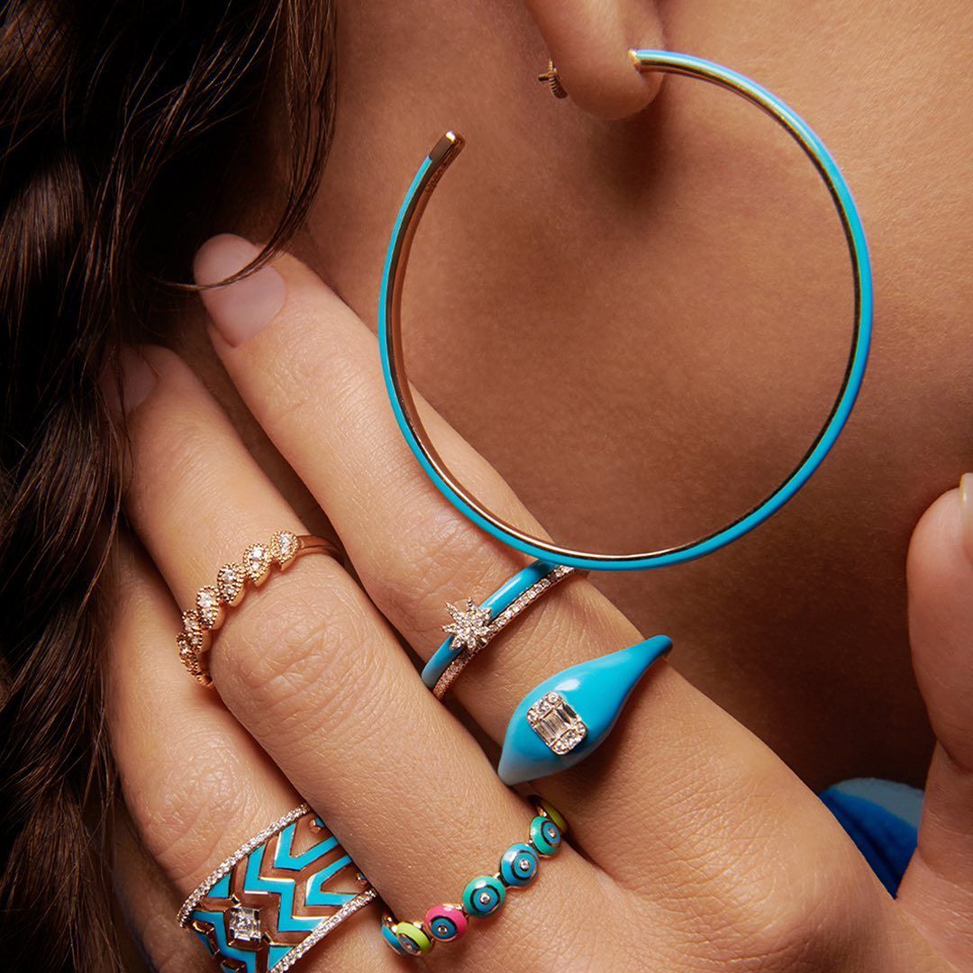 Main de femme avec des anneaux colorés et une grande boucle d'oreille créole turquoise.