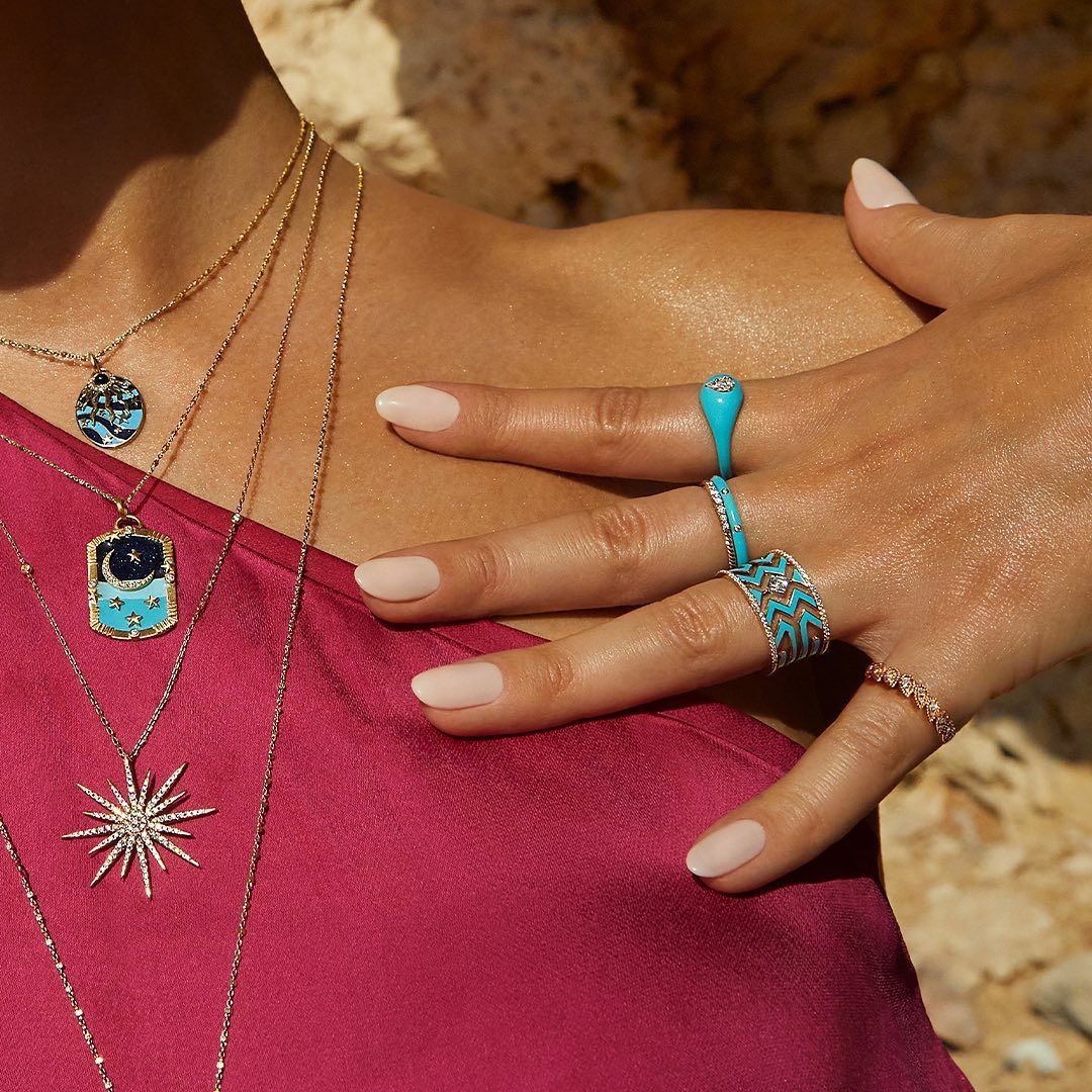 Main de femme avec bagues et colliers sur un haut rose. Bagues turquoise et divers colliers en or.