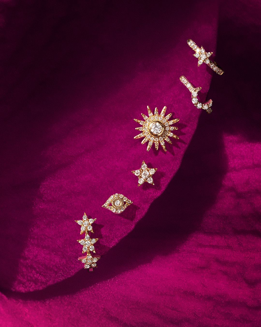 Parure de bijoux sur tissu magenta, comprenant des boucles d'oreilles en forme d'étoile, de soleil et de croissant de lune avec diamants.
