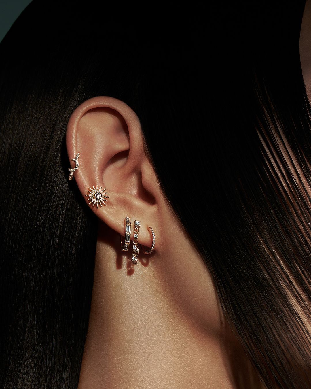 Oreille ornée de multiples boucles d'oreilles : en forme de soleil, créoles et clous. Des cheveux foncés encadrent l'oreille.