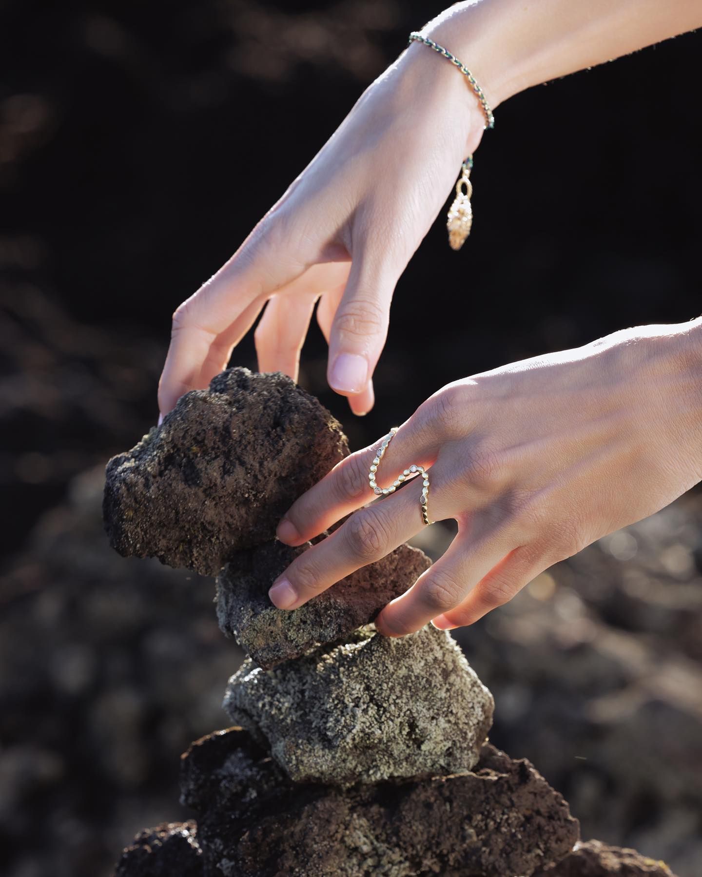 Des mains empilent soigneusement des pierres sombres et brutes, portant des bijoux, sur un fond flou.