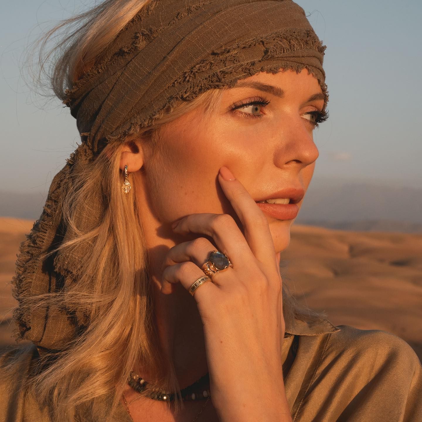 Femme blonde dans le désert, foulard sur la tête, touchant sa joue. Bijoux, lumière dorée.