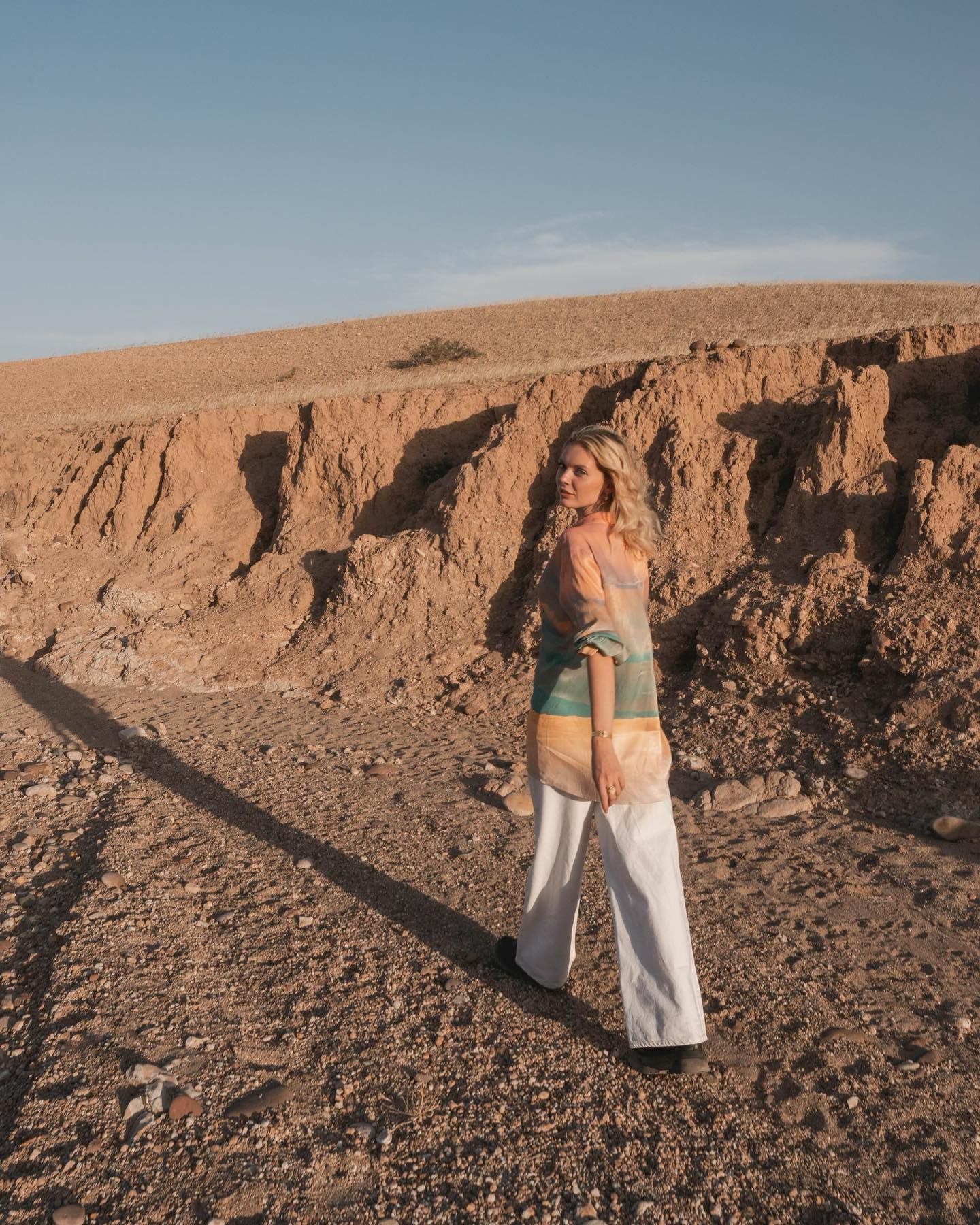 Une femme blonde en haut coloré et pantalon blanc regarde en arrière, debout sur un chemin de terre. Paysage désertique.