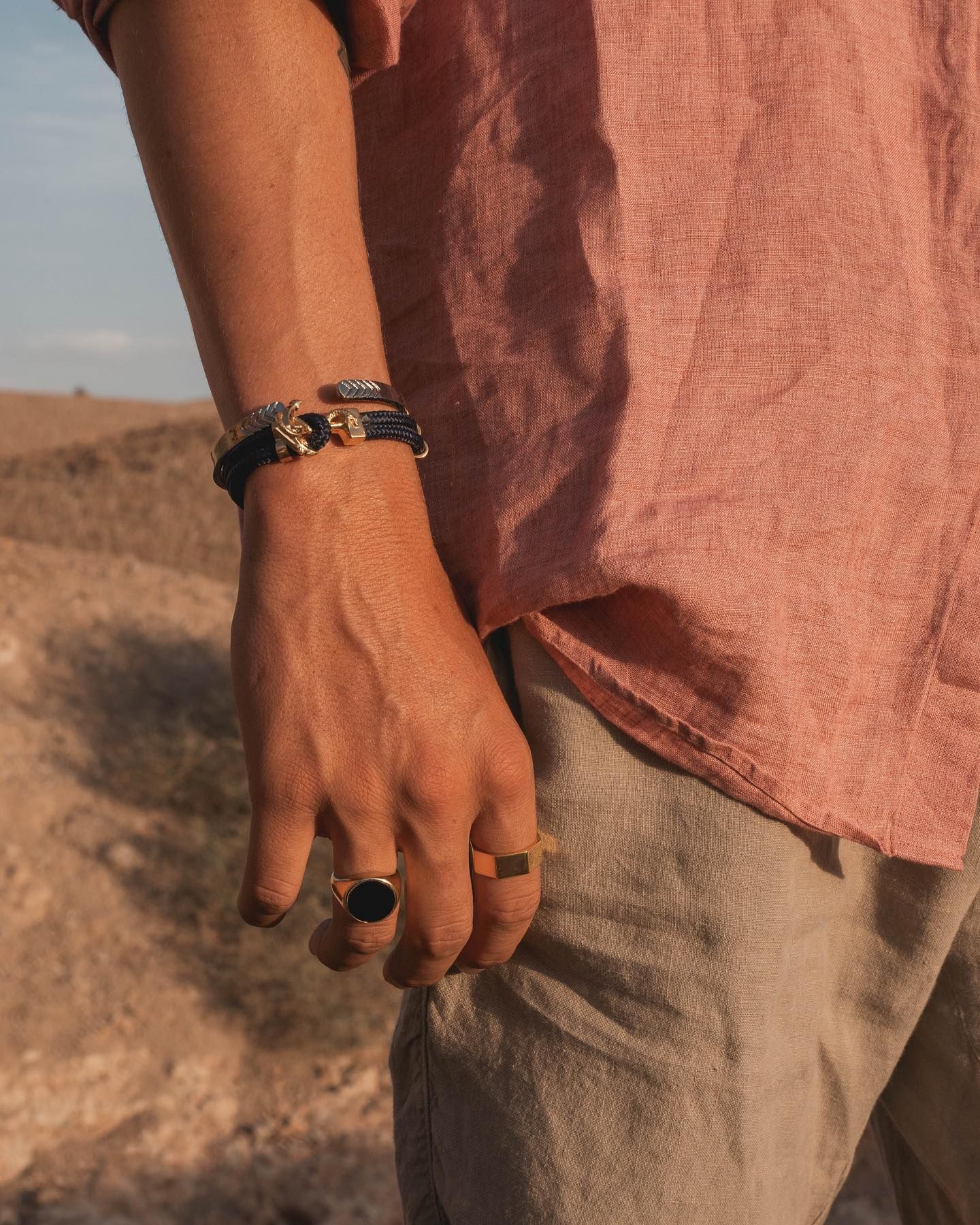 Bras d'homme avec bracelet et bagues, portant une chemise rose et un pantalon gris, dans un décor extérieur.
