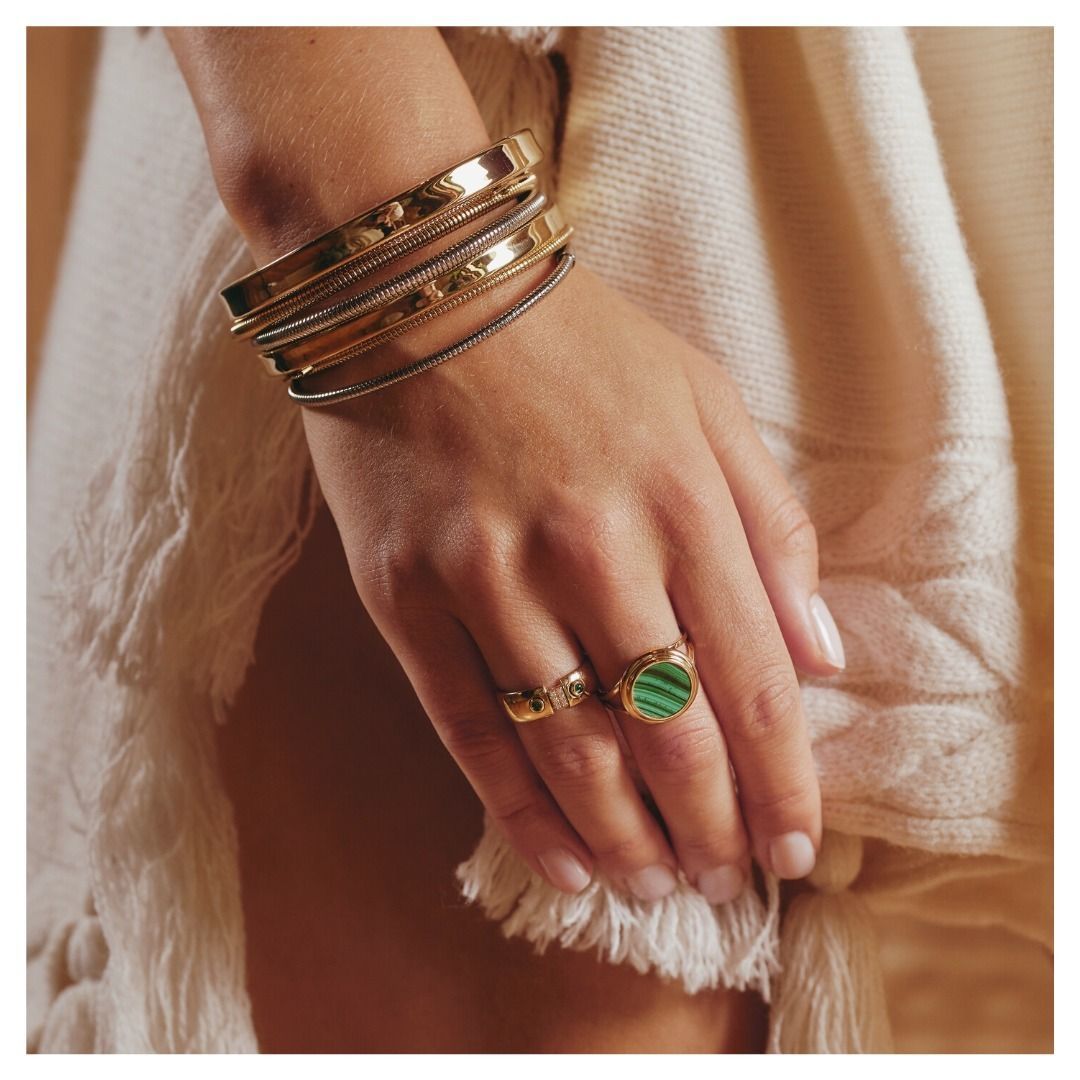 Main de femme ornée de bracelets et de bagues en or, dont une avec malachite verte, reposant sur un tissu crème.