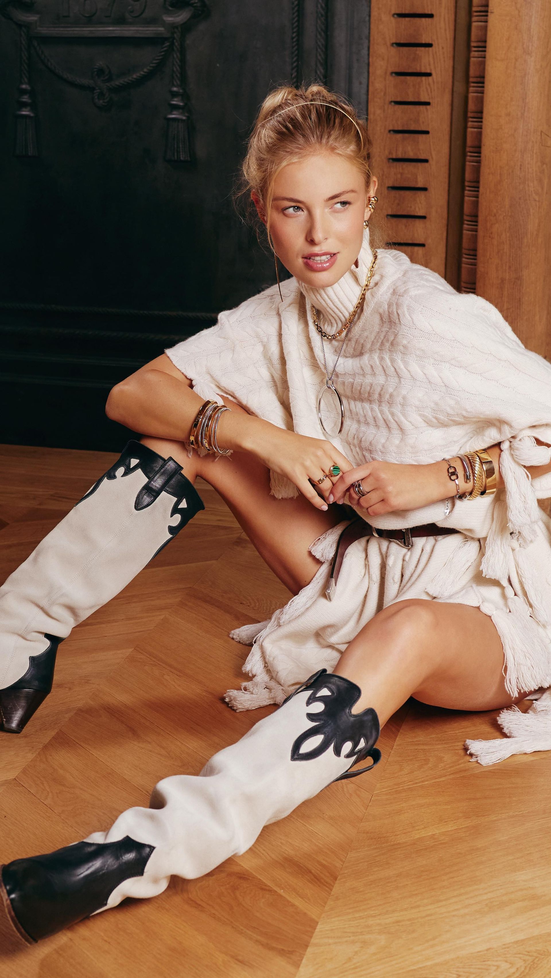 Femme en pull et jupe de couleur crème, bottes de cow-boy noires et blanches, assise sur un plancher en bois.