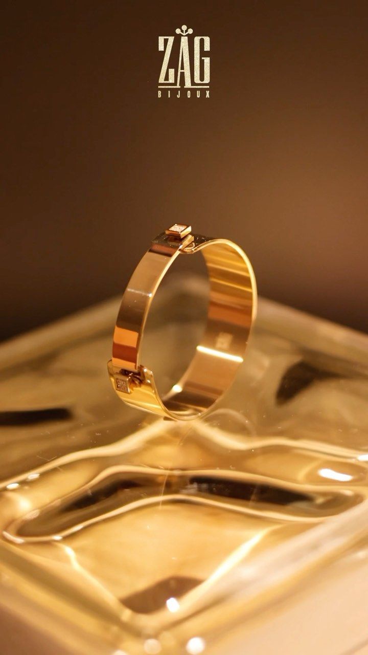 Créole en or suspendue au-dessus d'une surface réfléchissante, éclairée d'une lumière dorée. Logo ZAG Bijoux en haut.