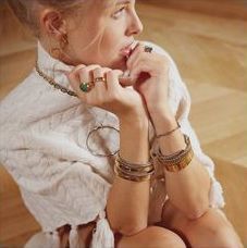 Femme en pull blanc, plusieurs bagues et bracelets, posée les mains jointes, à l'intérieur.