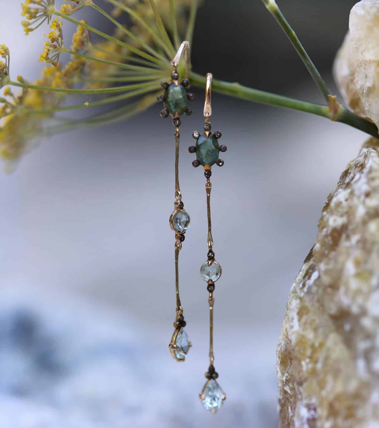 Boucles d'oreilles en or avec pierres vertes et bleues, suspendues à une fleur jaune.