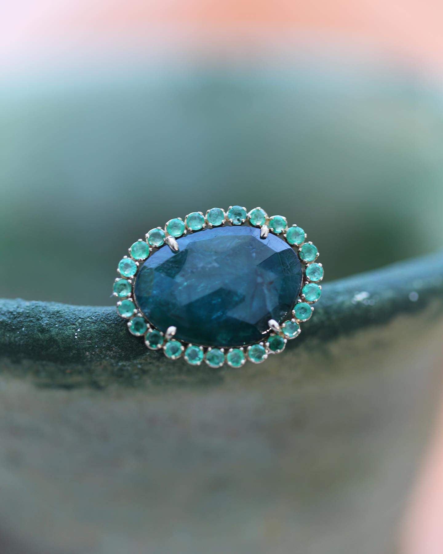 Bague émeraude ovale, grande pierre centrale, entourée d'émeraudes plus petites, reposant sur une surface bleu sarcelle.