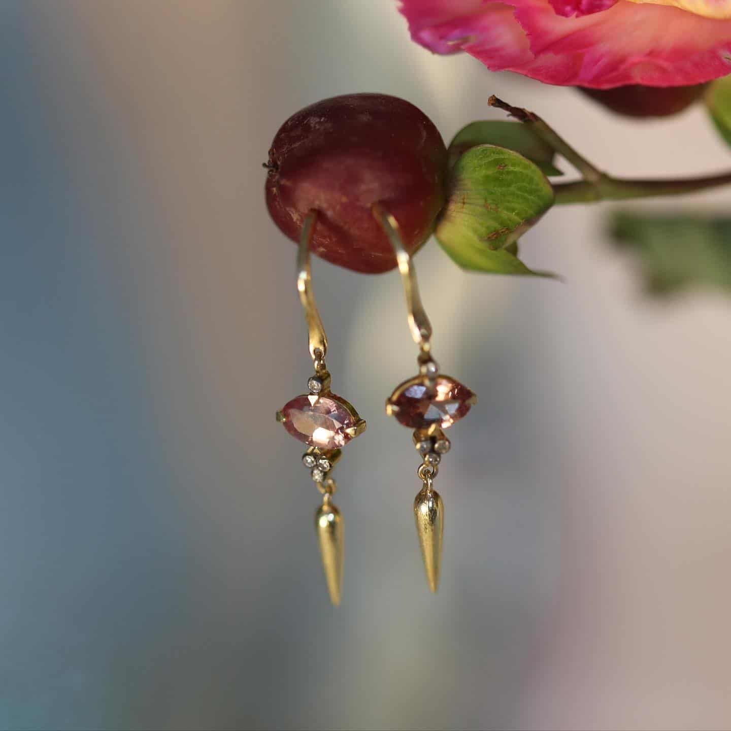 Boucles d'oreilles en or avec une pierre précieuse mauve, suspendue à côté d'une baie et d'une fleur rose.