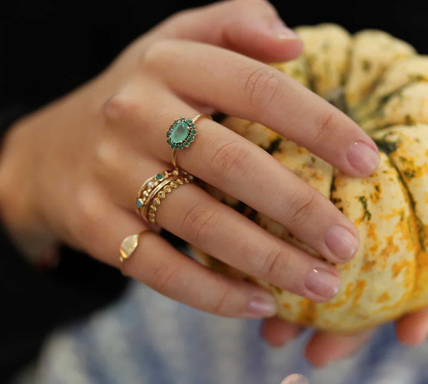 Main tenant une petite citrouille jaune et verte, ornée de plusieurs anneaux d'or ornés de pierres précieuses vertes.