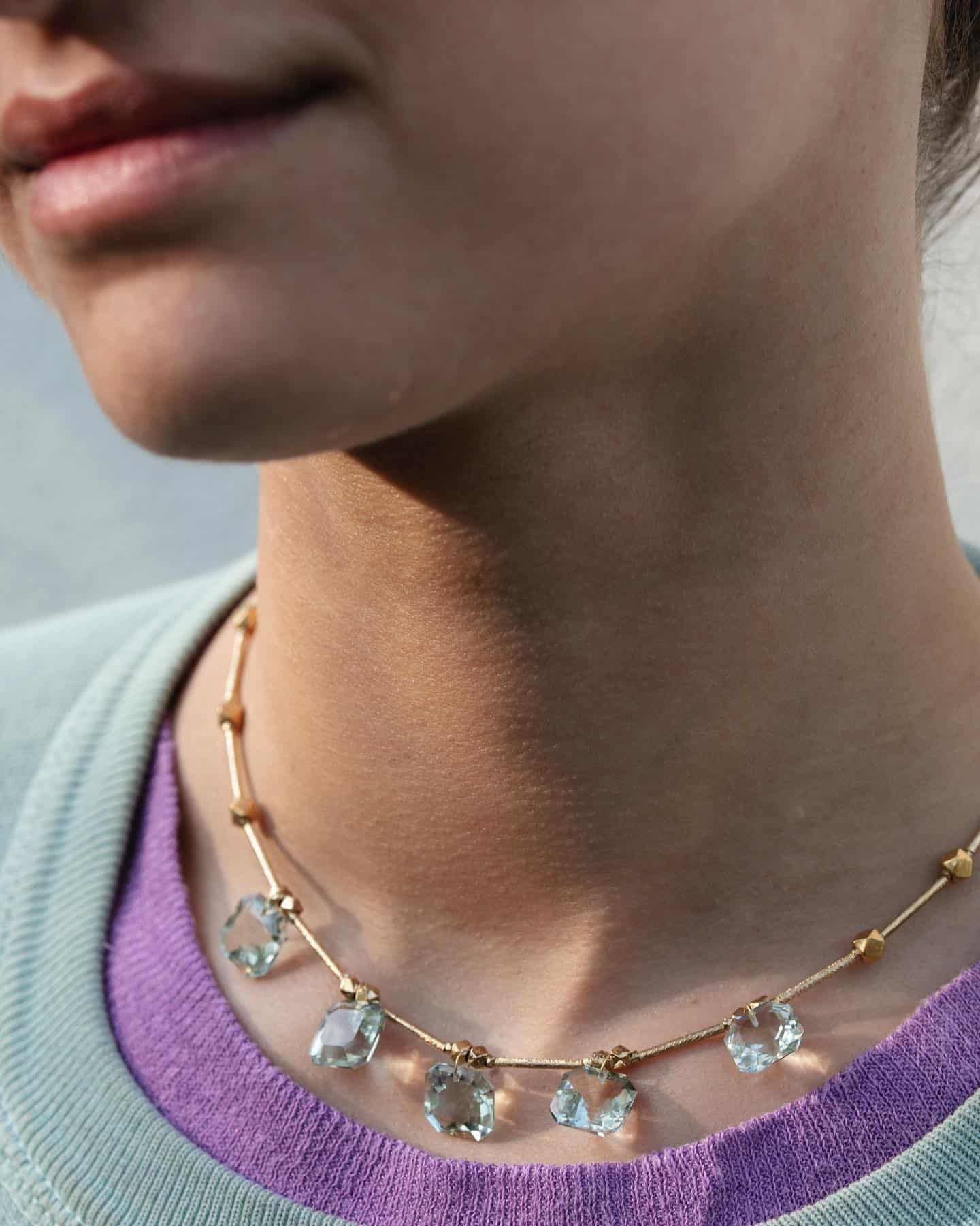Personne portant un collier en or avec des pendentifs en pierres précieuses vertes. Pull bleu clair sur une chemise violette.