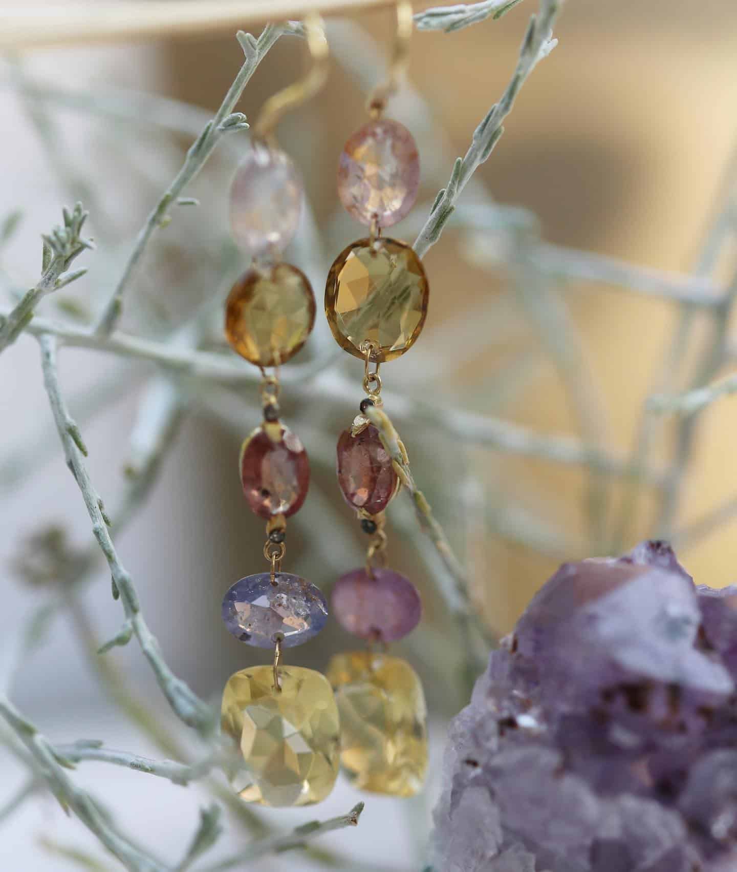 Boucles d'oreilles pendantes en or avec pierres précieuses colorées, disposées sur des brindilles à côté d'un cristal violet.